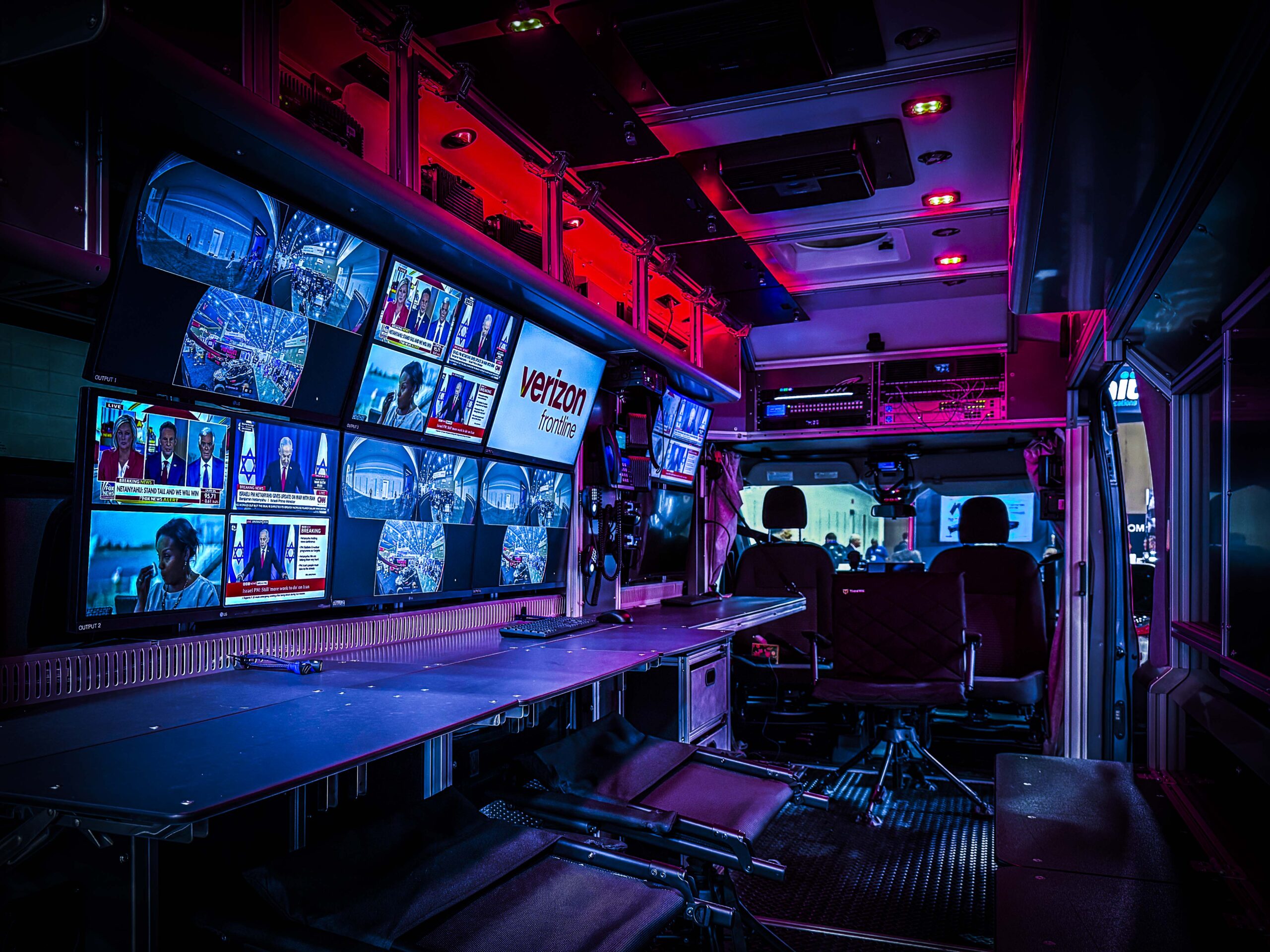 Inside of a mobile command center, illuminated by red and blue ambient lighting. A long dark desk runs along the left side, facing a large multi-screen video wall displaying various news broadcasts and surveillance feeds.